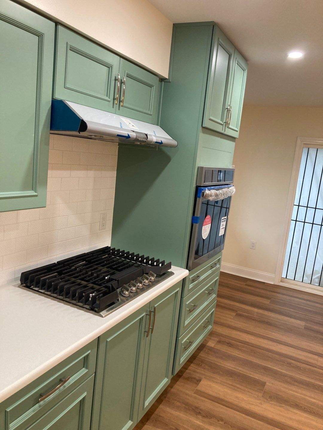 A gas cooktop under a cabinet-front hood, flanked by a stainless wall oven and sage-green upper cabinets.